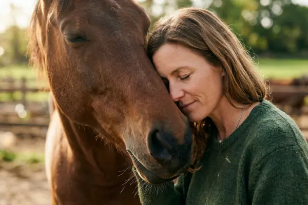 Nahaufnahme einer tiefen, vertrauensvollen Verbindung: Eine glückliche Frau umarmt ihr braunes Pferd auf der Weide, die Augen geschlossen. Symbolbild für die emotionale Beziehung zwischen Reiter und Pferd.