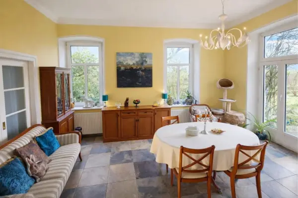 Ein geräumiges, helles Esszimmer mit gelben Wänden und großen weißen Fenstern, die den Blick nach draußen ins Grüne freigeben. In der Mitte steht ein runder Tisch mit einer weißen Tischdecke, um den vier Holzstühle platziert sind. An den Wänden stehen antike Holzmöbel, darunter eine Kommode und eine Vitrine, auf denen Dekorationsgegenstände arrangiert sind. Links im Bild befindet sich ein gestreiftes Sofa mit blauen Kissen und rechts steht ein Kratzbaum für Katzen. Der Bodenbelag besteht aus großen, unregelmäßigen quadratischen Fliesen in Grau- und Brauntönen. An der Decke hängt ein geschwungener weißer Kronleuchter.