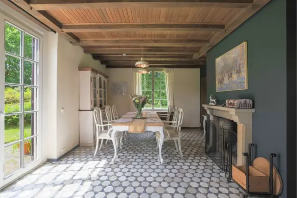Ein stilvolles Esszimmer im Landhausstil mit sichtbarer Holzbalkendecke und einem geometrisch gemusterten Fliesenboden. In der Raummitte steht ein langer Esstisch mit heller Holzplatte und geschwungenen weißen Beinen, umstellt von passenden weißen Stühlen. An der dunkelgrünen Akzentwand auf der rechten Seite befindet sich ein offener Kamin mit Schutzgitter und einem gerahmten Pferdebild darüber. Eine große, bodentiefe weiße Sprossentür auf der linken Seite lässt viel Tageslicht herein und gibt den Blick in den grünen Garten frei