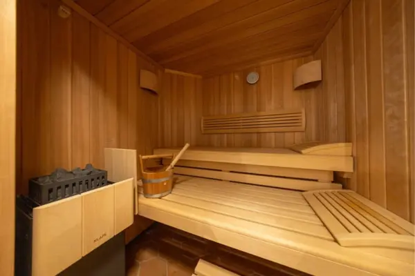 Innenansicht einer modernen Holzsauna mit mehrstufigen Liegebänken aus hellem Holz. Im linken Vordergrund befindet sich ein elektrischer Saunaofen mit Saunasteinen, während auf der unteren Liegefläche ein hölzerner Aufgusseimer samt Kelle sowie eine ergonomische Holz-Kopfstütze bereitstehen. Die warm ausgeleuchteten Wände sind rundum vertäfelt und mit eckigen Wandlampen, einem runden Messinstrument sowie horizontalen Lüftungsschlitzen ausgestattet.