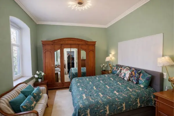 Ein gemütlich eingerichtetes Schlafzimmer mit dezent grün gestrichenen Wänden. Zentraler Blickfang ist ein großes Doppelbett, das mit einer auffälligen, blau-grünen Tagesdecke mit Tiermotiven bezogen ist. Mehrere bunte Kissen mit Blumenmustern zieren das Kopfende des Bettes. Links an der Wand steht ein massiver, antiker Kleiderschrank aus Holz mit Spiegeltüren. Vor dem Fenster, auf der linken Seite des Zimmers, lädt ein kleines, gestreiftes Sofa mit farblich zur Bettdecke passenden Kissen zum Verweilen ein. An der Wand über dem Kopfende des Bettes hängt eine große, weiße Tafel oder Leinwand, und auf den Nachttischen stehen dekorative Lampen.