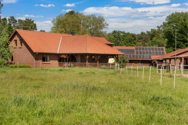 Blick über eine weite, grüne Weide mit Koppelzaun auf ein ländliches Hofensemble mit rot verklinkerten Gebäuden und roten Ziegeldächern. Links steht das Wohnhaus mit einer gemütlichen, überdachten Holzveranda, während das mittlere Nebengebäude mit einer Photovoltaikanlage auf dem Dach ausgestattet ist. Rechts schließen sich Stallungen an, vor denen ein dunkles Pferd am Paddockzaun steht, eingerahmt von dichtem Baumbestand unter einem blauen Himmel mit Schönwetterwolken.