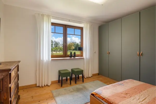Ein helles Schlafzimmer mit warmem Holzdielenboden, weißen Wänden und einem großen Holzfenster, das den Blick ins Grüne und auf die umliegenden Weiden freigibt. An der rechten Wand erstreckt sich ein raumhoher, moderner Kleiderschrank in mattem Graugrün. Unter dem von weißen Vorhängen eingerahmten Fenster stehen zwei olivgrüne Polsterhocker, während links eine antike, kunstvoll verzierte Holzkommode platziert ist. Im Vordergrund ist das Fußende eines Bettes mit einer warm gemusterten Tagesdecke auf einem hellen Vorleger zu sehen.