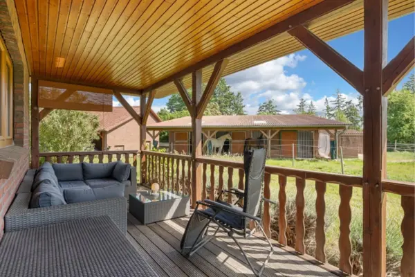 Blick von einer überdachten Holzveranda mit rustikalem Holzgeländer und holzvertäfelter Decke direkt auf das ländliche Grundstück. Auf der Terrasse lädt eine gemütliche, dunkelgraue Lounge-Ecke aus Polyrattan samt kleinem Glastisch und einem schwarzen Liegestuhl zum Verweilen ein. Direkt gegenüber, jenseits einer Grünfläche, befindet sich ein langgestrecktes Stallgebäude mit offenen Paddockboxen, in denen ein Schimmel und ein dunkles Pferd stehen. Bäume, ein weiteres Backsteingebäude auf der linken Seite und ein strahlend blauer Himmel mit weißen Wolken im Hintergrund unterstreichen die idyllische Hofatmosphäre.