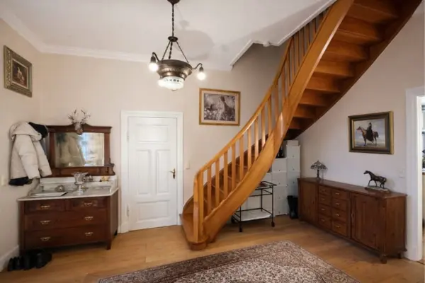 Ein geräumiger Flur mit hellen Wänden und einem warmen Holzboden, auf dem ein großer, gemusterter Teppich liegt. Im Zentrum des Raumes führt eine geschwungene Holztreppe mit einem stabilen Geländer nach oben. Links von der Treppe befindet sich eine weiße Tür, daneben steht eine antike Kommode mit Spiegel, an der eine weiße Jacke hängt. Rechts von der Treppe steht ein langes, niedriges Sideboard aus Holz, auf dem eine kleine Pferdeskulptur platziert ist. An den Wänden hängen mehrere gerahmte Bilder, und von der Decke leuchtet eine auffällige, schmiedeeiserne Lampe.