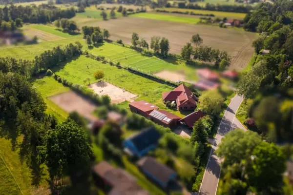Eine Luftaufnahme im Miniatur-Effekt (Tilt-Shift-Optik), die ein idyllisches Hofensemble mit roten Ziegeldächern und angrenzenden Pferdeweiden zeigt. Auf dem Anwesen befinden sich das Wohnhaus, Nebengebäude mit einer Photovoltaikanlage sowie zwei eingezäunte Sandreitplätze inmitten weitläufiger grüner Wiesenflächen. Rechts führt eine schmale Landstraße am Grundstück vorbei, das ringsum von dichten Baumreihen und landwirtschaftlichen Feldern eingerahmt wird.
