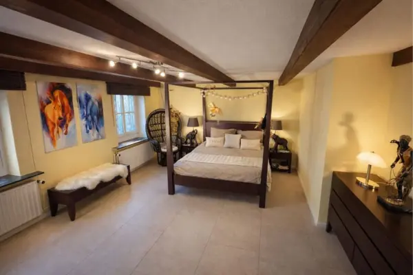 Ein geräumiges Schlafzimmer mit hellen Fliesen, cremefarbenen Wänden und einer Decke mit markanten dunklen Holzbalken. In der Mitte steht ein großes Himmelbett aus dunklem Holz, das mit beigefarbener Bettwäsche bezogen und mit einer Lichterkette am Kopfteil dekoriert ist. Rechts daneben befindet sich eine dunkle Holzkommode mit einer Tischlampe und einer afrikanisch anmutenden Skulptur. Links vom Bett steht ein auffälliger, hoher Korbsessel, und vor den zwei Fenstern an der linken Wand ist eine dunkle Holzbank mit einer weißen Fellauflage platziert. Über der Bank hängen zwei farbenfrohe Gemälde, die Pferde in kräftigen Orange- und Blautönen zeigen