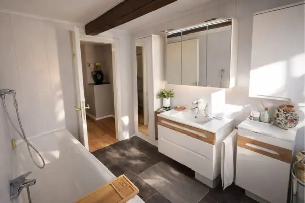 Ein helles, sonnendurchflutetes Badezimmer mit weißen, holzvertäfelten Wänden und einem markanten, dunklen Holzbalken an der Decke. Der Boden ist mit großen, dunklen Fliesen ausgelegt, auf denen ein kleiner grauer Vorleger liegt. Im linken Vordergrund befindet sich eine weiße Badewanne mit Duscharmatur und einer hölzernen Ablage. Auf der rechten Seite steht ein weißer Waschtisch mit hellem Holzakzent an der großen Schublade, darüber hängt ein dreitüriger, beleuchteter Spiegelschrank. Daneben ergänzen ein schmaler Unterschrank im gleichen Design sowie ein Hängeschrank die Ausstattung. Durch die geöffnete weiße Tür im Hintergrund ist ein angrenzender Raum mit Holzboden und einer dekorativen Vase zu sehen.
