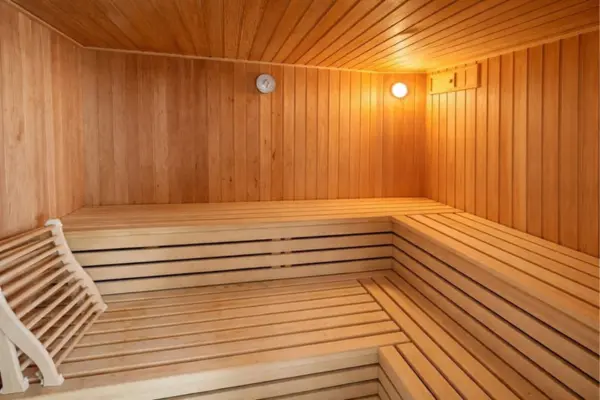 Innenansicht einer Sauna mit warmtoniger Holzvertäfelung an Wänden und Decke. Über Eck angeordnete, mehrstufige Sitz- und Liegebänke aus hellem Holz bieten Platz zum Entspannen. Im linken Vordergrund liegt eine geschwungene Kopfstütze aus Holzlatten auf der Bank. An der Rückwand befinden sich ein rundes Messinstrument sowie eine warm leuchtende Saunalampe in der oberen Ecke.