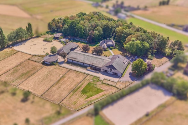 Tilt-Shift-Luftbild eines idyllischen Reitguts mit einem großen Wohnhaus, angrenzenden Ställen und mehreren abgezäunten Pferdewiesen.