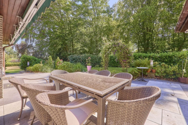 Eine gepflasterte Terrasse mit einer Rattan-Essgruppe, bestehend aus einem Tisch und Stühlen mit Kissen. Im Hintergrund ein üppiger grüner Garten mit Bäumen und einem Blumenbogen. Links die Hausfassade mit Backsteinen und einer Markise.