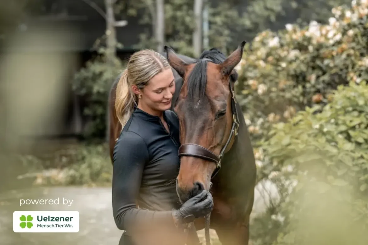 Ein Farbfoto, auf dem eine blonde Frau mit Pferdeschwanz in schwarzer Reitkleidung zärtlich ihren Kopf an ein braunes Pferd mit schwarzer Mähne schmiegt. Sie trägt einen Reithandschuh und hält ein schwarzes Seil. Der Hintergrund besteht aus Büschen mit weißen Blüten. Im linken unteren Bereich ist ein grünes Logo mit der Aufschrift „Uelzener Mensch. Tier. Wir.“ und dem Text „powered by“ zu sehen. Der Blick der Frau ist gesenkt, ein leichtes Lächeln liegt auf ihrem Gesicht.