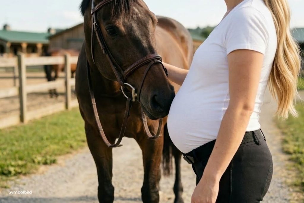 Eine realistische Nahaufnahme im Querformat auf einem Reiterhof. Eine schwangere Frau mit langen blonden Haaren steht im Profil rechts im Bild. Sie trägt ein weißes T-Shirt, das ihren deutlich gewölbten Bauch betont, und eine schwarze Reithose. Ein braunes Pferd mit Lederzaumzeug steht links vor ihr und stupst mit seiner weichen Nase sanft und behutsam gegen ihren Bauch. Der Fokus liegt auf der liebevollen Interaktion zwischen Tier und Mensch; das Gesicht der Frau ist nicht vollständig zu sehen. Im Hintergrund erkennt man einen Reitplatz mit Holzzäunen und Stallgebäude unter einem hellen Himmel.