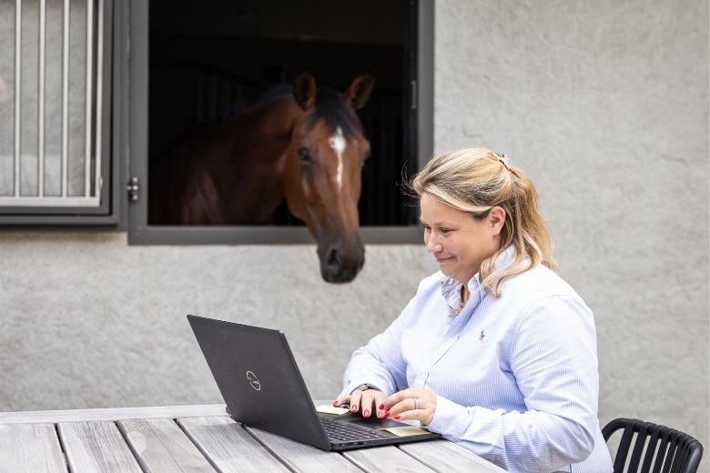 Wie geht richtiges Stallmanagement? Frau lernt am Laptop, im Hintergrund steht Pferd