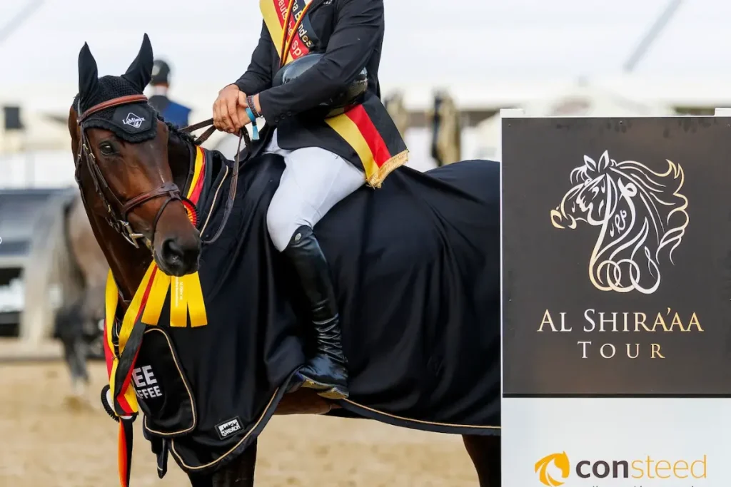 Ein siegreiches Pferd mit Reiter in schwarz-rot-goldener Bundeschampionats-Schärpe neben einer Stele mit dem verschnörkelten Pferdekopf-Logo der Al Shira'aa Tour. Foto: sportfotos-lafrentz.de