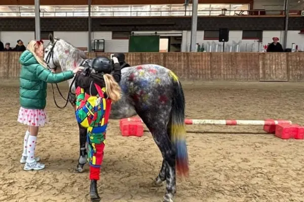 In einer Reithalle steht ein Apfelschimmel, dessen Fell mit vielen bunten Farbtupfern bemalt ist und dessen Schweif in mehreren Farben (Gelb, Blau, Rot) leuchtet. Links am Kopf des Pferdes steht eine Person mit grüner Daunenjacke und Rock. Eine zweite Person in einem bunten Patchwork-Kostüm und Reithelm steht seitlich am Pferd. Im Hintergrund sind der Sandboden der Reithalle, die hölzerne Bande sowie ein rot-weißes Bodenhindernis zu sehen.