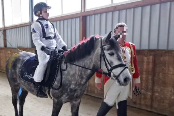 In einer Reithalle sitzt ein Kind, verkleidet als Astronaut in einem weißen NASA-Overall und Helm, auf einem gesprenkelten Apfelschimmel-Pferd. Das Pferd trägt eine dunkle Schabracke mit der Aufschrift „Sea Biscuit“ und hat rote Verzierungen in der Mähne sowie einen gelben Farbfleck an der Seite. Rechts neben dem Pferd geht ein Erwachsener, gekleidet in eine rote britische Offiziersuniform mit Goldbesatz, und führt das Pferd an der Trense. Der Hintergrund besteht aus Holzpaneelen und einer Metallwand mit Fenstern.