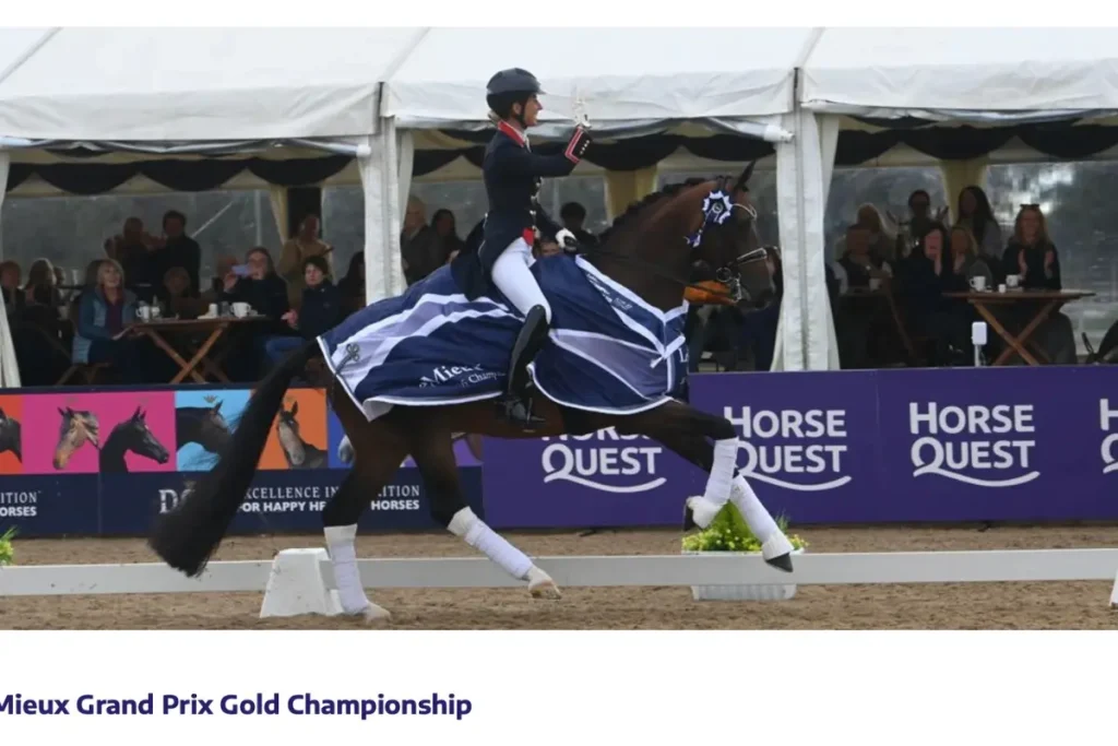 Ein Foto zeigt Charlotte Dujardin auf dem dunkelbraunen Pferd Brave Heart während einer Ehrenrunde nach dem Gewinn der britischen Dressur-Nationals im Grand Prix. Charlotte Dujardin, in formeller Reitkleidung, reitet mit einer Hand am Zügel und der anderen erhoben, um dem applaudierenden Publikum zuzuwinken. Brave Heart trägt eine dunkelblaue Siegerdecke mit weißen Akzenten, weiße Bandagen und eine Siegerschleife am Zaumzeug. Im Hintergrund sind Zuschauer unter einem Zelt und Bandenwerbung mit Texten wie "HORSE QUEST" zu sehen. Der Text "Mieux Grand Prix Gold Championship" ist unten im Bild zu sehen. Foto: British Dressage/Kevin Sparrow.