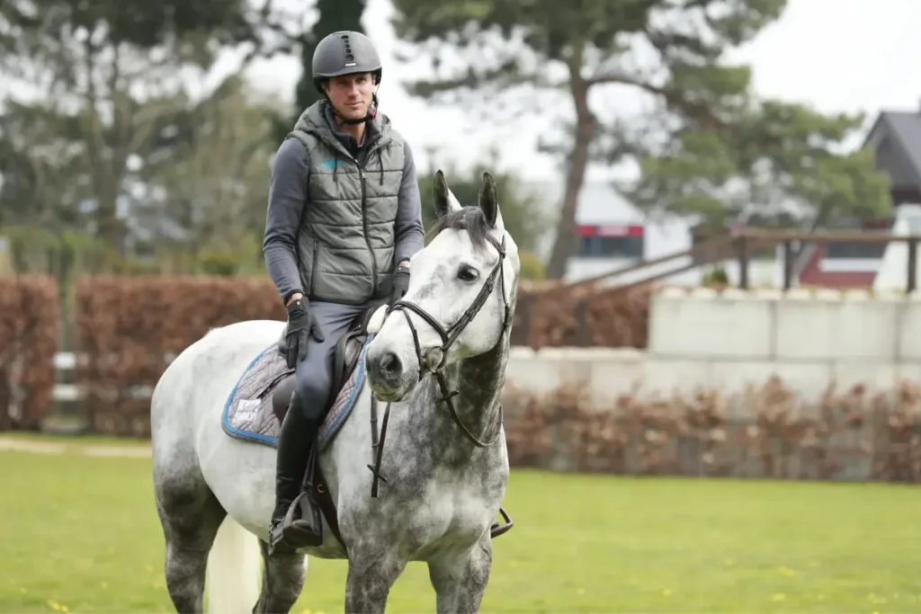 Christian Kukuk sitzt auf seinem grauen Schimmel-Pferd Checker auf einem Reitplatz im Freien. Er trägt einen Reithelm und eine graue gesteppte Weste.