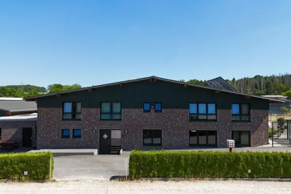 Außenansicht des modernen, zweigeschossigen Haupt- und Funktionsgebäudes der Reitanlage in Heimbach am Nationalpark Eifel. Die Fassade kombiniert dunkle Klinker-Backsteine im Erdgeschoss mit einer passenden, dunklen Holzverkleidung im Giebelbereich unter einem flachen Satteldach. Vor dem Gebäude mit seinen dunklen Fensterrahmen liegt ein gepflegter, gepflasterter Hofbereich, der im Vordergrund von einer niedrig geschnittenen, grünen Hecke eingefasst wird. Die Aufnahme entstand an einem sonnigen Tag unter strahlend blauem Himmel.