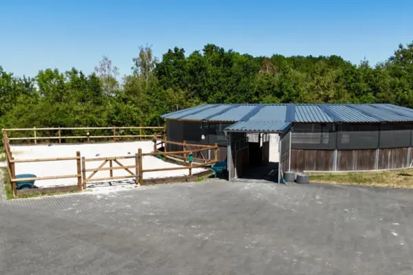 Außenansicht der Trainingsanlagen auf dem Gelände der Reitanlage in Heimbach am Nationalpark Eifel. Rechts befindet sich eine moderne, überdachte Führanlage mit dunklem Trapezblechdach, Holzverkleidung und Windschutznetz. Links daneben schließt sich ein eingezäunter Sandpaddock mit hellem Boden und Holzgatter an. Der asphaltierte Hofbereich erstreckt sich im Vordergrund, während das Areal im Hintergrund an eine grüne Baumkulisse unter strahlend blauem Himmel grenzt.
