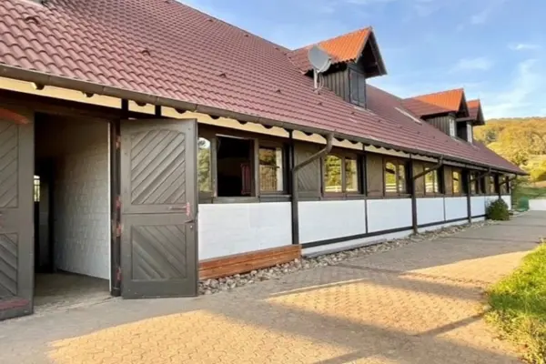 Außenansicht eines langgestreckten Stallgebäudes im Fachwerkstil mit geöffnetem Holztor und gepflastertem Weg.