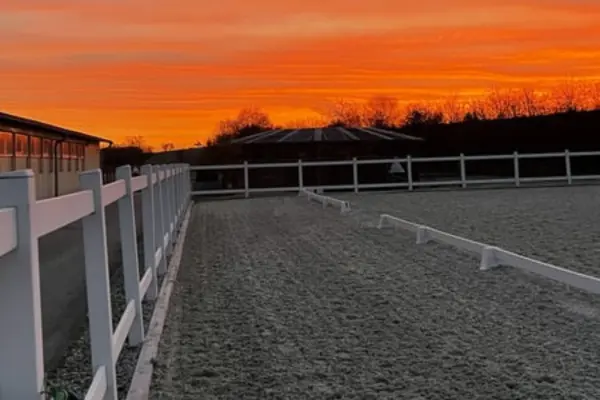 Ein Reitplatz im Freien mit einem weißen Holzzaun und niedrigen Begrenzungselementen vor dem Hintergrund einer runden Reithalle und einem dramatischen, intensiv orange-roten Sonnenuntergangshimmel.