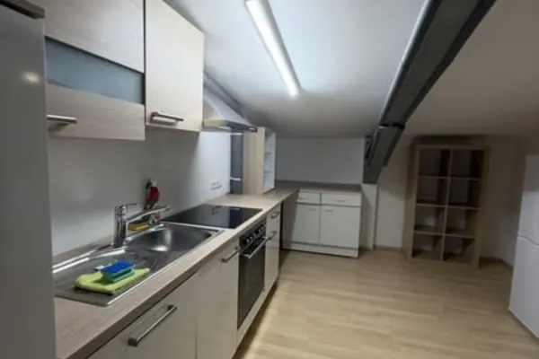 Blick in eine funktionale, helle Küchenzeile unter einer Dachschräge. Die Küche ist mit Edelstahlspüle, Cerankochfeld, Backofen und Oberschränken ausgestattet. Im hinteren Bereich befindet sich ein Unterschrank an der Wand, rechts steht ein offenes Standregal auf hellem Holzboden.