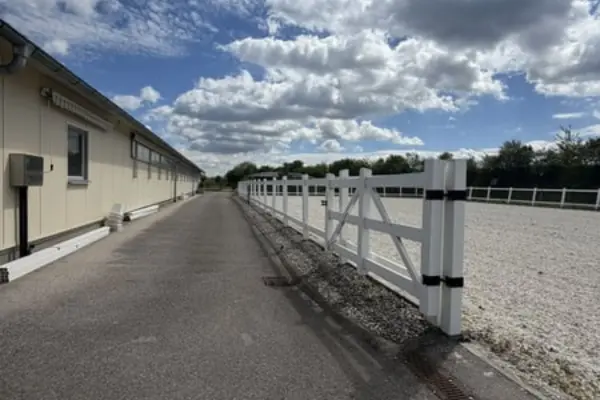 Außenaufnahme einer Reitanlage. Ein asphaltierter Weg verläuft links, parallel zu einem langen, weißen Pferdezaun, der einen sandigen Reitplatz auf der rechten Seite begrenzt. Im Hintergrund ist ein langes, helles Stallgebäude zu sehen, umgeben von Bäumen unter einem dramatisch bewölkten blauen Himmel.