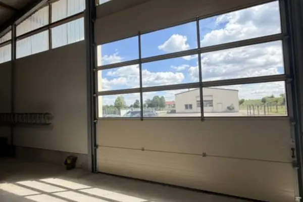 Blick aus dem Inneren einer Halle durch ein Sektionaltor mit verglasten Elementen. Durch die Fensterscheiben sieht man auf das Außengelände mit einem weiteren flachen Betriebsgebäude und Bäumen unter blauem Himmel mit leichten Wolken.