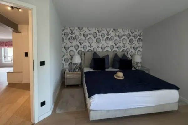 Modern gestaltetes Schlafzimmer mit einem Doppelbett, das mit einer dunkelblauen Decke und dunklen Kissen hergerichtet ist. Auf dem Bett liegt ein Strohhut. Die Kopfwand ist mit einer grau-weißen Muster-Tapete im Heißluftballon-Design versehen, seitlich stehen zwei Nachttische mit passenden Lampen.