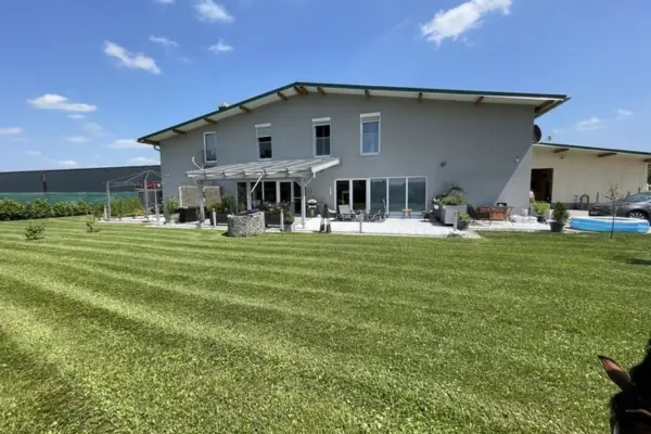 Rückansicht eines modernen, zweigeschossigen grauen Gebäudes mit flachem Satteldach und großer Terrasse. Im Vordergrund erstreckt sich ein weitläufiger, streifenförmig frisch gemähter Rasen unter sonnigem, blauem Himmel.
