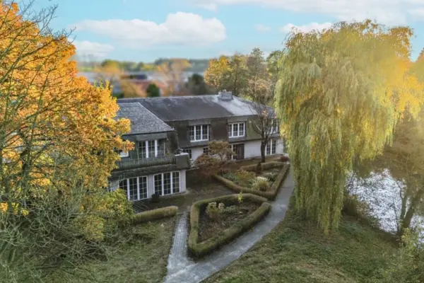 Eine Luftaufnahme eines eleganten, zweistöckigen Hauses mit dunklem Dach und heller Fassade, eingebettet in eine herbstliche Gartenlandschaft an einem Gewässer. Ein leuchtend orangefarbener Herbstbaum auf der linken Seite und eine große Weide auf der rechten Seite, die von warmem Sonnenlicht beleuchtet wird, rahmen das Haus ein. Ein gepflasterter Weg führt durch formal gestaltete, geometrische Heckenbeete zu einer Terrasse im Erdgeschoss.
