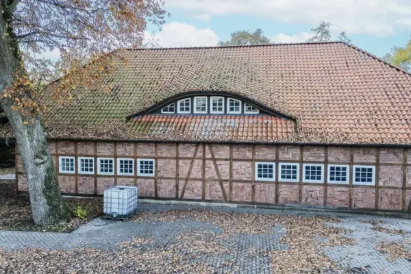 Historisches Fachwerkgebäude aus Backstein mit rotem Ziegeldach und einer charakteristischen Fledermausgaube. Die Fassade ist mit einer Reihe weißer Sprossenfenster versehen, während auf dem mit Herbstlaub bedeckten Pflasterhof davor ein weißer IBC-Wassertank steht.