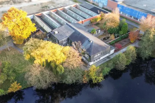 Luftaufnahme eines weitläufigen Anwesens an einem dunklen Gewässer im Herbst. Das Gebäude mit dunklem Dach grenzt an einen großen Hallenkomplex mit mehreren verglasten Sheddächern. Bunt gefärbte herbstliche Bäume und eine Gartenanlage umschließen das Grundstück.