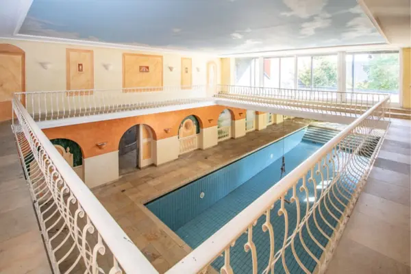 Blick von einer Empore mit weißem Ziergeländer hinab auf ein großes, blau gefliestes Hallenbad. Der Raum ist im mediterranen Stil mit terrakotta- und beigefarbenen Wandbögen gestaltet, während die Decke mit einer Wolkenhimmel-Bemalung versehen ist. Eine große Fensterfront lässt Tageslicht herein.