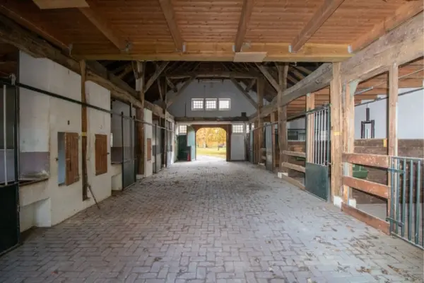 Innenansicht einer gepflasterten Stallgasse mit freiliegenden Holzbalken und urigem Dachgebälk. Links und rechts befinden sich Pferdeboxen mit dunkelgrünen Metall- und Holzabtrennungen. Am Ende der Gasse gibt ein geöffnetes Scheunentor unter kleinen Sprossenfenstern den Blick auf eine herbstliche Außenanlage frei.