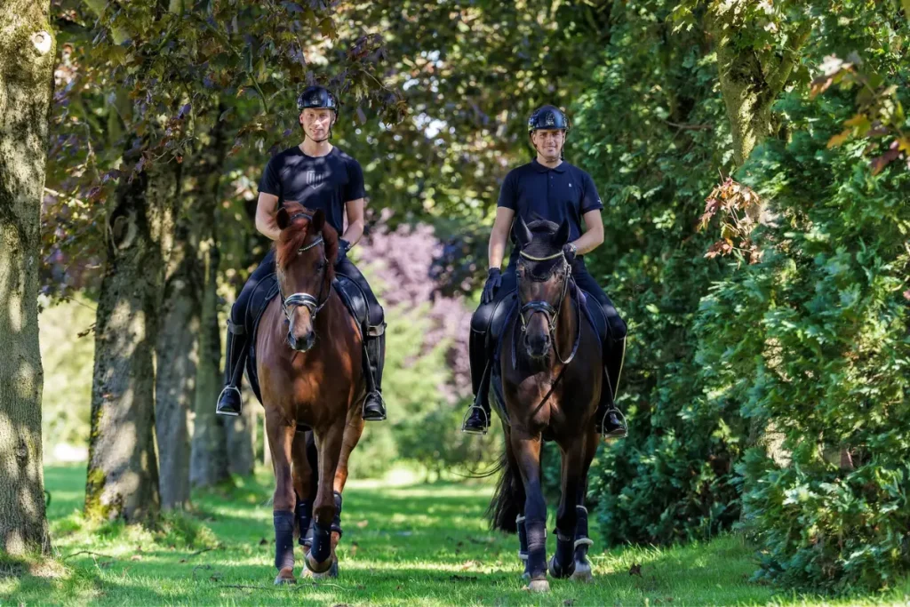 Zwei Männer, Frederic Wandres und Lars Ligus, reiten auf zwei Pferden nebeneinander auf einem grasbewachsenen, von Bäumen gesäumten Weg. Frederic Wandres links sitzt auf einem Fuchs und trägt ein schwarzes T-Shirt und einen Helm. Lars Ligus rechts sitzt auf einem Dunkelbraunen und trägt ein schwarzes Poloshirt und einen Helm. Beide Pferde tragen Sättel und Zäume, und das rechte Pferd hat eine Fliegenhaube auf. Sie schauen in die Kamera, während sie durch eine natürliche, schattige Reitweg-Allee an einem sonnigen Tag reiten.