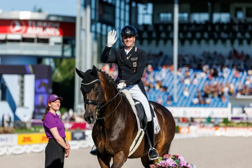 Der deutsche Dressurreiter Frederik Wandres sitzt auf seinem braunen Wallach Bluetooth OLD und winkt lächelnd dem Publikum im Stadion zu. Er trägt einen dunklen Turnierfrack mit Deutschland-Emblem, weiße Reithosen, einen Helm und weiße Handschuhe. Links neben dem Pferd steht eine Turnierhelferin in einem lila Poloshirt vor gefüllten Zuschauerrängen.