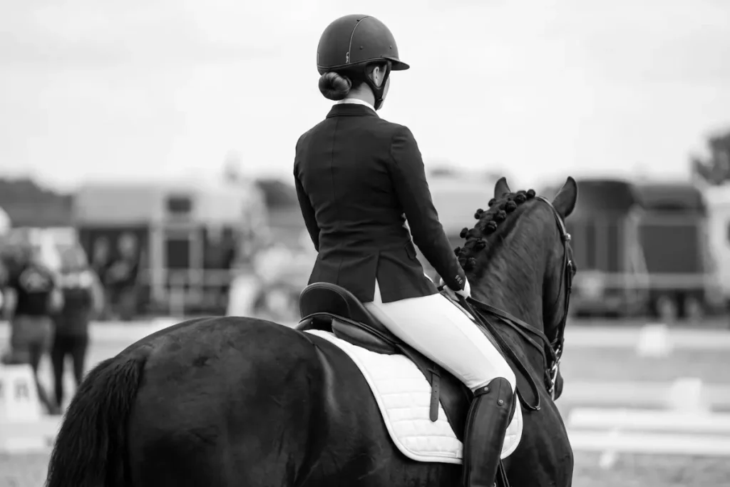 Fotografie aus der Perspektive leicht schräg hinter einem Dressurreiter-Paar auf einem Reitplatz. Eine Reiterin mit hochgestecktem Dutt im Nacken und schwarzem Reithelm sitzt auf einem tiefschwarzen Pferd mit ordentlich eingeflochtener Mähne. Sie trägt ein dunkelblaues Turniersakko, eine weiße Reithose, schwarze Lederstiefel und nutzt eine weiße Schabracke unter dem Sattel. Im Hintergrund befindet sich eine voll besetzte, überdachte Zuschauertribüne aus Holz sowie weitere Zuschauer, die am Rand des Dressurvierecks stehen. Das Bild ist scharf auf Pferd und Reiterin fokussiert, während der Hintergrund in einer leichten Unschärfe liegt.