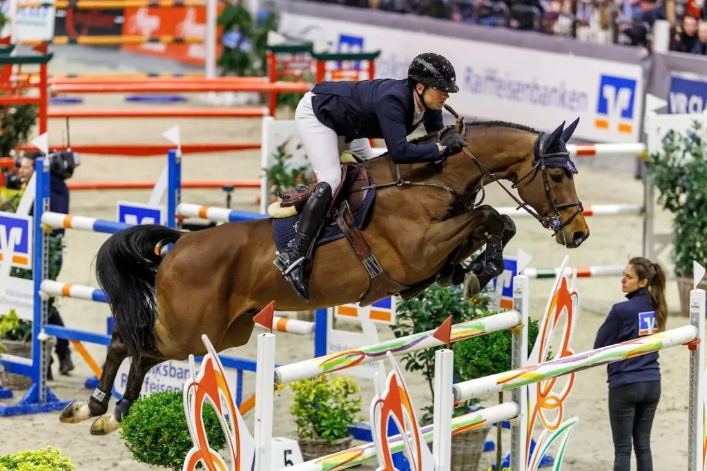 Ein professionelles Foto des Holsteiner Hengstes United Way, einem braunen Warmblut, das von Lucas Wenz geritten wird. Sie überwinden gerade ein großes, dekoratives Springhindernis mit floralen und schmetterlingsartigen Mustern bei den VR Classics in Neumünster 2026. Der Hintergrund zeigt die Hallenreitbahn mit Tribünen und Werbebannern für Volksbanken Raiffeisenbanken. Eine Frau steht beobachtend an der Seite. (Foto: sportfotos-lafrentz.de)