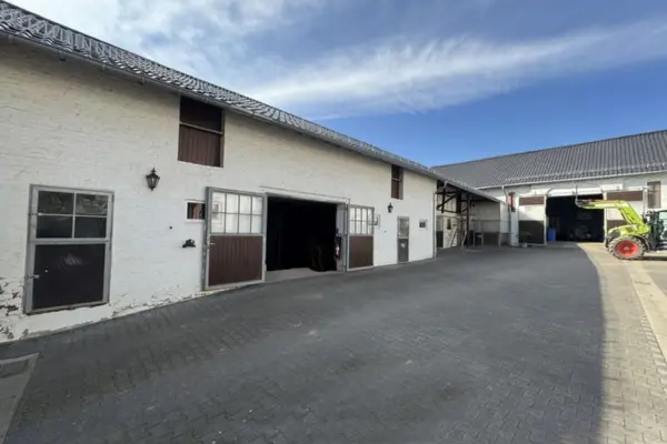 Innenhof des historischen, renovierten Vierkanthofs mit Reitanlage in Kreuzau. Im Zentrum und auf der linken Seite steht ein großes, zweigeschossiges Stallgebäude mit weiß verputzter Backsteinfassade und dunklem Ziegeldach, das im Erdgeschoss mehrere geteilte Boxentüren und Ladeluken im Obergeschoss besitzt. Eine große, geöffnete Tordurchfahrt führt in das Gebäude. Der weitläufige, gepflasterte Innenhof führt im rechten Hintergrund zu einem weiteren, rechtwinklig anschließenden Wirtschafts- und Stalltrakt, vor dem ein grüner Traktor mit Frontlader abgestellt ist. Die Aufnahme wurde an einem leicht bewölkten Tag bei hellem Tageslicht aufgenommen.