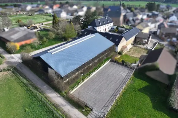 Luftaufnahme aus der Vogelperspektive der Reitanlage des historischen Vierkanthofs in Kreuzau. Im Vordergrund steht eine große Reithalle mit blau-grauem Metalldach und durchgehendem Lichtband am First, direkt daneben befindet sich ein rechteckiger, eingezäunter Außenreitplatz mit hellem Sandboden. Im Hintergrund schließt sich das weiß verputzte Hofensemble mit dunklem Dach an, eingebettet in grüne Wiesen- und Koppelfflächen am Rande der Ortschaft.
