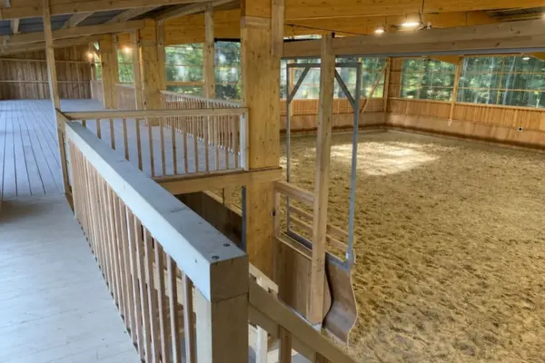 Innenansicht einer leeren, modernen Reithalle aus Holz, aufgenommen von einer erhöhten Plattform. Im Vordergrund befindet sich ein Aussichtssteg mit Holzgeländer. Die weitläufige Reitfläche ist mit hellem Sand bedeckt. Große Fensterfronten im Hintergrund bieten einen Ausblick auf einen Wald. Die Decke wird von massiven Holzbalken getragen.