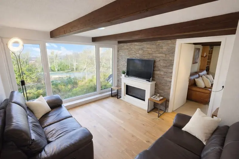 Ein helles Wohnzimmer mit großen Panoramafenstern, die den Blick auf eine Landschaft mit Bäumen und einen Fluss oder See freigeben. Zwei braune Ledersofas mit hellen Kissen stehen sich gegenüber. Der Boden ist mit hellem Holzparkett ausgelegt. An der Wand gegenüber den Fenstern befindet sich eine Steinverkleidung mit einem weißen Kamin, auf dem ein Flachbildfernseher steht. Die weiße Decke ist mit dunklen Holzbalken durchzogen. Durch eine geöffnete Tür auf der rechten Seite ist ein weiterer Raum mit einem ähnlichen braunen Ledersofa zu sehen. In der Ecke links am Fenster steht eine moderne Stehlampe mit einem leuchtenden Ring.