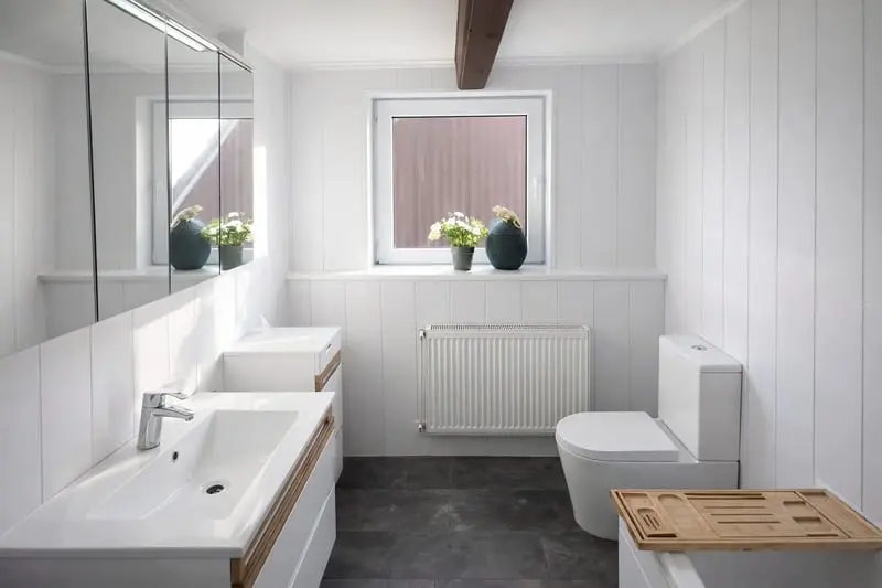 Ein helles, modern gestaltetes Badezimmer mit dunkelgrauen Bodenfliesen, weiß vertäfelten Wänden und einem dunklen Deckenbalken aus Holz. Links befindet sich ein weißer Waschtisch mit Holzakzent an der oberen Kante und einem großen, dreitürigen Spiegelschrank darüber. An der Stirnseite fällt Tageslicht durch ein Fenster, auf dessen Fensterbank Topfpflanzen stehen, direkt über einem weißen Heizkörper. Rechts stehen eine moderne weiße Toilette sowie im Vordergrund eine Badewanne mit einer hölzernen Wannenablage.