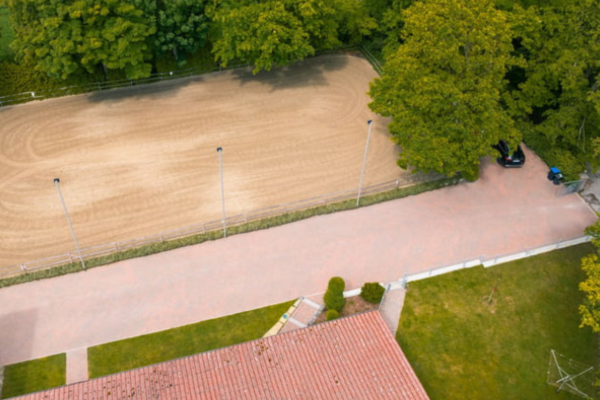 Vogelperspektive einer beleuchteten Sand-Reitarena, umgeben von dichten grünen Bäumen, mit angrenzendem Gebäude und Auffahrt.