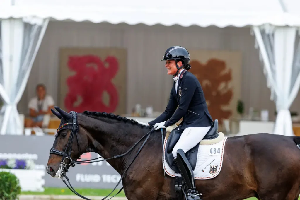 Ein Foto aus der Seitenansicht zeigt die deutsche Paralympics-Medaillengewinnerin Isabell Nowak auf ihrem dunkelbraunen Dressurpferd. Sie sitzt aufrecht, lächelt und trägt einen schwarzen Reithelm mit Glitzersteinen, eine schwarze Reitjacke mit deutschen Team-Insignien (Deutschland-Flagge, Bundesadler) und weiße Reithosen. Das Pferd hat eine geflochtene Mähne und trägt eine weiße Schabracke mit der Startnummer "484", dem Text "WALDHAUSEN" und einem großen Bundesadler. Der Hintergrund zeigt einen Reitplatz mit einem Zelt, in dem zwei große, abstrakte rote Logos und ein unscharfer Zuschauer zu sehen sind.