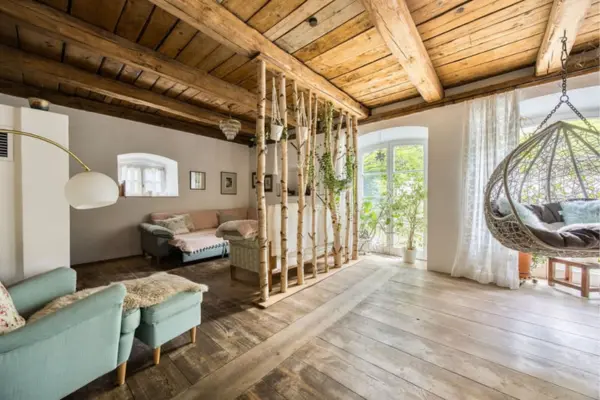 Einladender Wohnbereich im rustikal-modernen Stil mit naturbelassenem Dielenboden und einer Decke aus massiven Holzbalken. Ein Raumteiler aus vertikal aufgestellten Birkenstämmen trennt die Sitzecke mit Sofa vom übrigen Raum. Im Hintergrund fällt reichlich Tageslicht durch eine große Rundbogentür ins Zimmer.