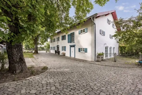 Seitenansicht eines weiß verputzten, traditionellen Landhauses mit dunkelblauen Klappläden. Im Vordergrund erstreckt sich eine weitläufige Zufahrt aus Kopfsteinpflaster, auf der linken Seite stehen große, schattenspendende Laubbäume.
